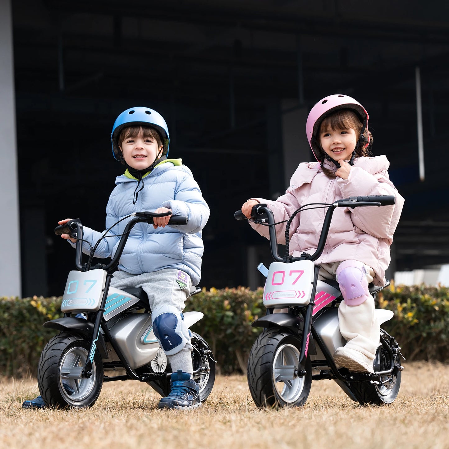 Deux enfants souriants sur des motos électriques 250W en extérieur, casques et protections genoux.