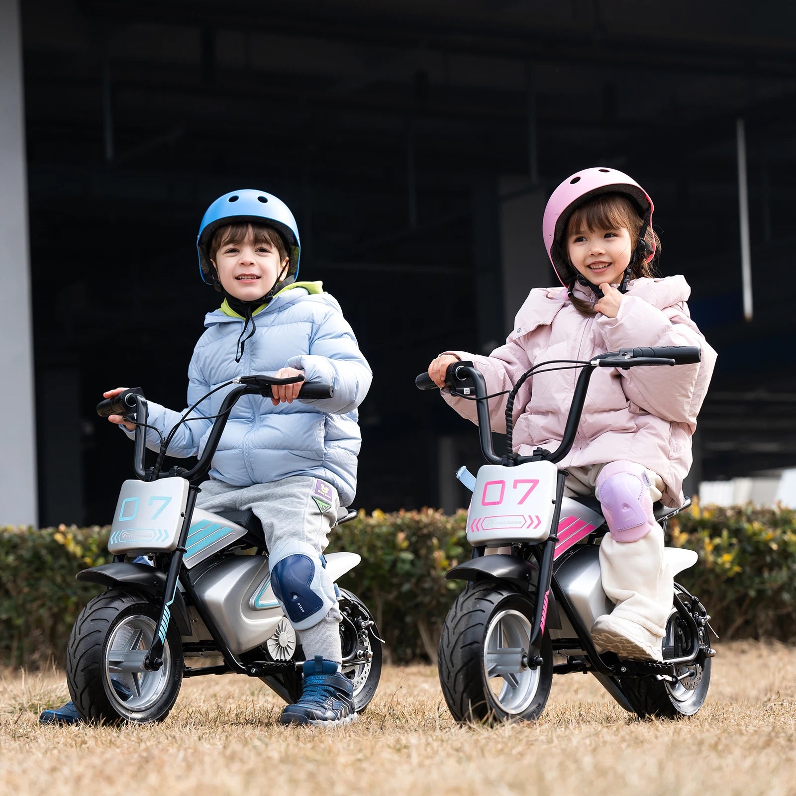 Deux enfants souriants sur des motos électriques 250W en extérieur, casques et protections genoux.